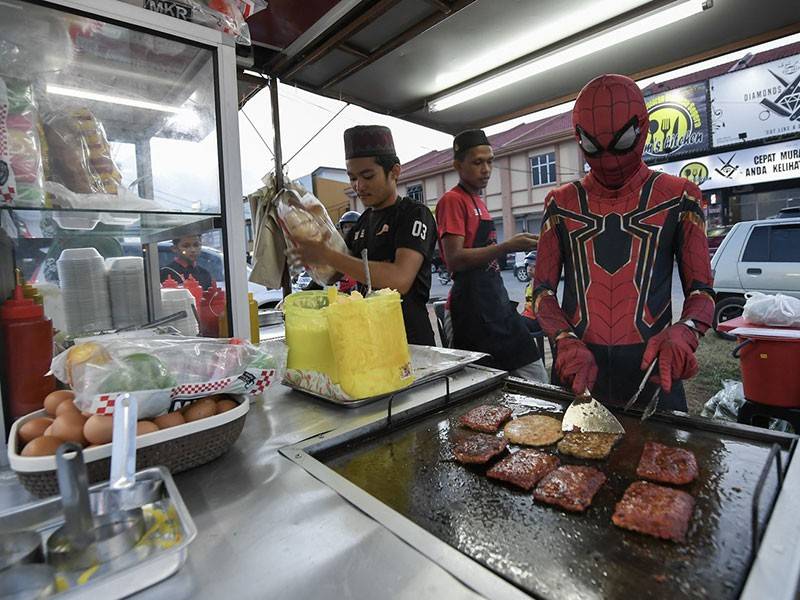 Fakhrul Fahmie Ishak, 18, (kanan) menggayakan kostum adiwira Spiderman turut membantu abangnya Ahmad Ridhuan, 22, (kiri) menyediakan pesanan burger di Kampung Raja baru-baru ini.- Foto Bernama