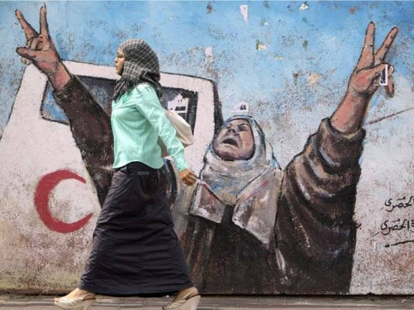 Seorang wanita Palestin berjalan di hadapan dinding dipenuhi lukisan mural di Gaza.
