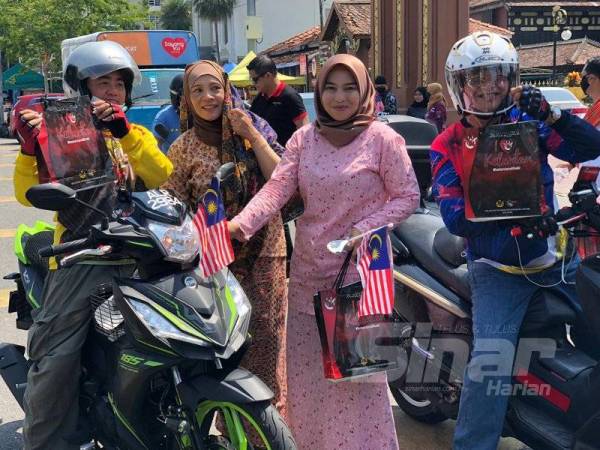 Raja Rusmin (dua dari kiri) memakai kain batik dan berkerudung mengagihkan bendera kepada pengguna jalan raya bersama Adila (dua dari kanan) berhampiran Padang Merdeka.