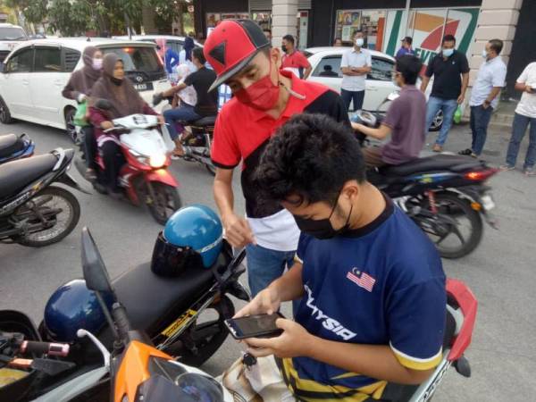 Pengerusi Skuad Sabil Kuala Terengganu, Armi Irzan Mohd (berdiri) membantu seorang pengunjung pasar di Bukit Bayas, Kuala Terengganu melengkapkan proses pendaftaran vaksin Covid-19.