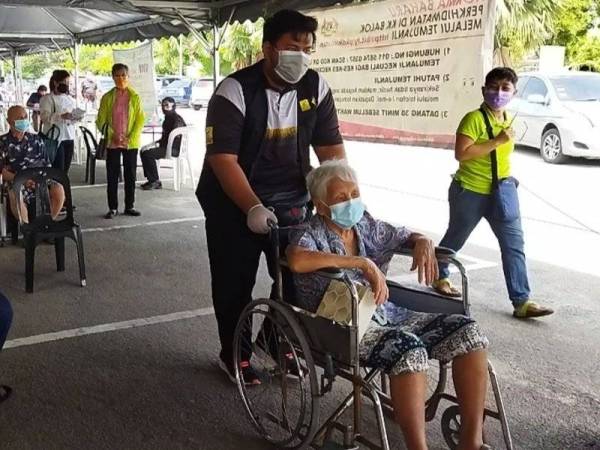 Sukarelawan Mampan membantu membawa warga emas menggunakan kerusi roda untuk masuk ke ruang yang disediakan bagi mendapatkan suntikan vaksin Covid-19 di Klinik Kesihatan Balok di sini, pada Sabtu.