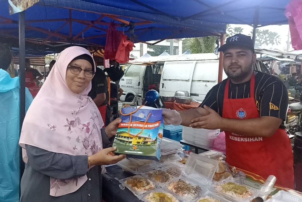 Dr Surinah (kiri) mengedarkan bahan pendidikan kesihatan kepada peniaga bazar Ramadan di Stadium Mini Pontian.