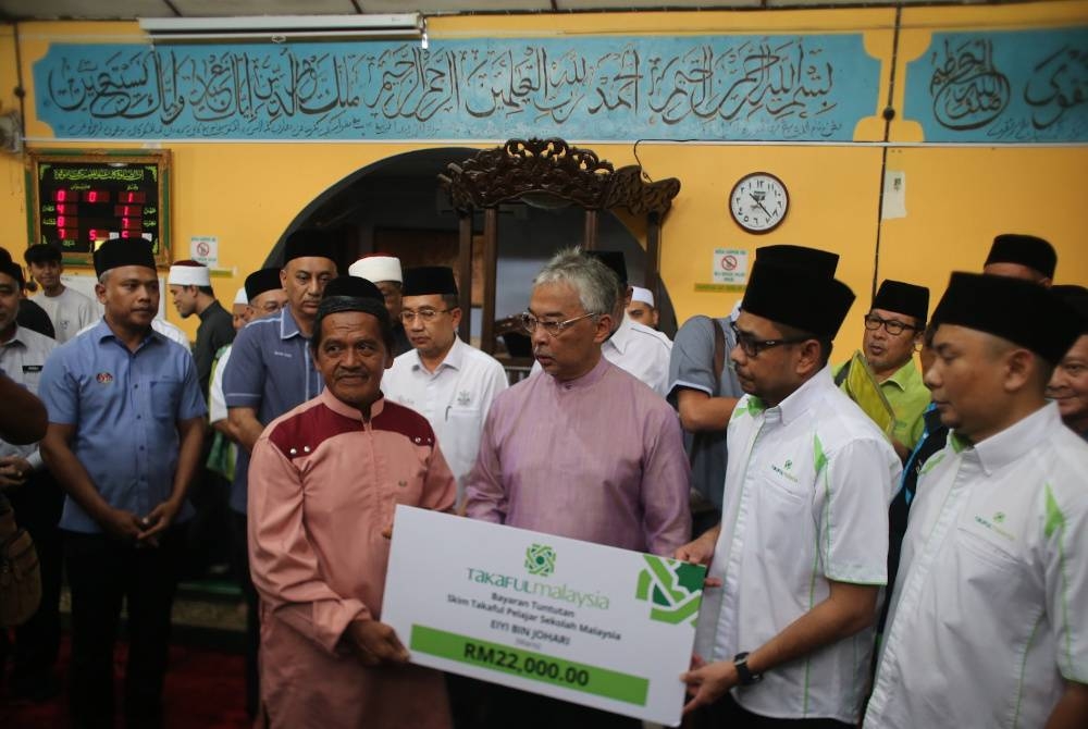 Eiyi (kiri) menerima replika cek daripada wakil Takaful disaksikan Al-Sultan Abdullah di Masjid Ubbudiah Felda Lepar Hilir 3 pada Isnin.