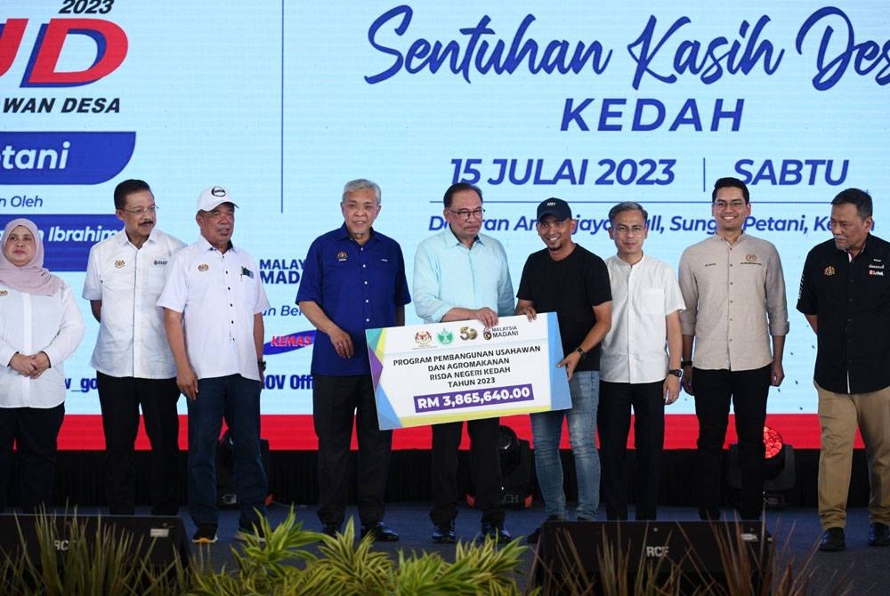 Anwar menyampaikan replika cek bantuan kepada wakil usahawan RISDA dari Kedah, Ainol Zulkarnain Zainal Abidin pada Program Sentuhan Kasih Desa Mini Karnival Usahawan Desa (KUD) Negeri Kedah di Dataran Amanjaya Mall, dekat sini pada Sabtu.