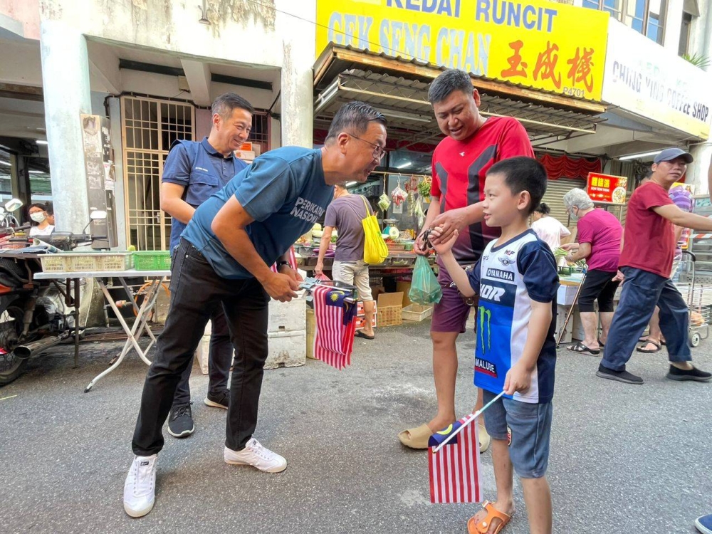 Dominic (kiri) memberi bendera kepada seorang kanak-kanak sempena Hari Malaysia di Pasar Awam Sungai Ara pada Sabtu.