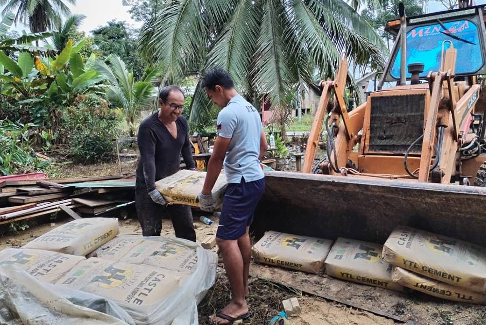 Raja Kamaruddin (kiri) memunggah simen yang digunakan untuk membina rumah baharu selepas ia musnah dibawa arus dalam banjir besar pada Disember tahun lalu.