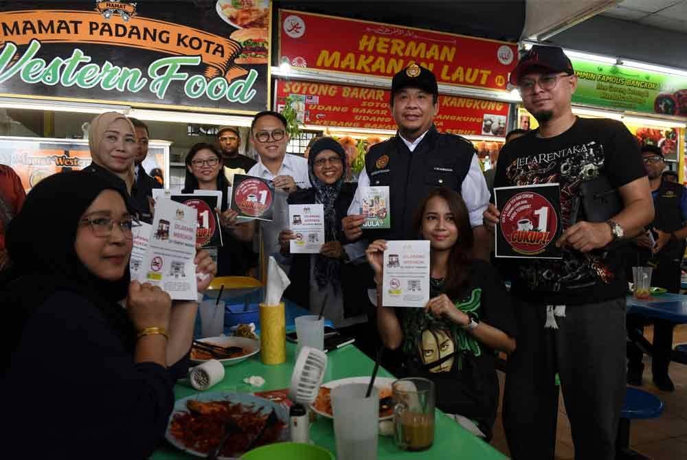 Lukanisman bersama orang awam menunjukkan risalah kurangkan gula dan dilarang merokok di tempat makan semasa membuat Program Walkabout Bebas di Kompleks Makanan Medan Renong, Padang Kota Lama pada Ahad. Foto Bernama